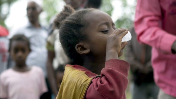 A little girl is drinking from a cup. In the background people are watching.