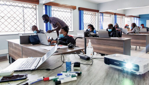 Students sit in front of laptops at desks in a classroom. In the foreground, a teacher stands next to a student, giving instruction.