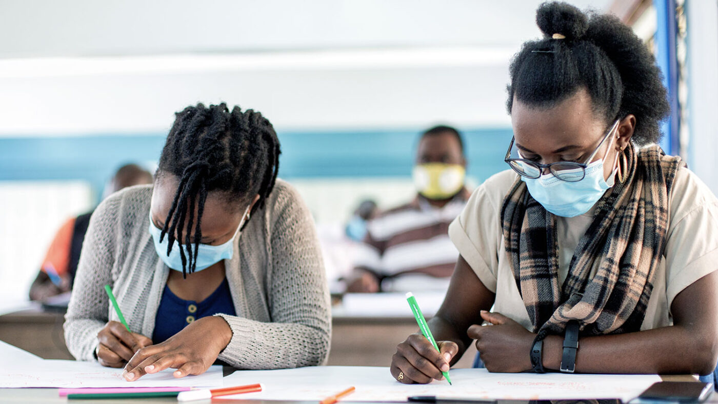 Two female students are sitting at a desk in a classroom, writing in notebooks. Shanice is sitting on the right.