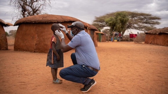 A young girl has her eyes checked for trachoma, with a rural village in Kenya in the background.