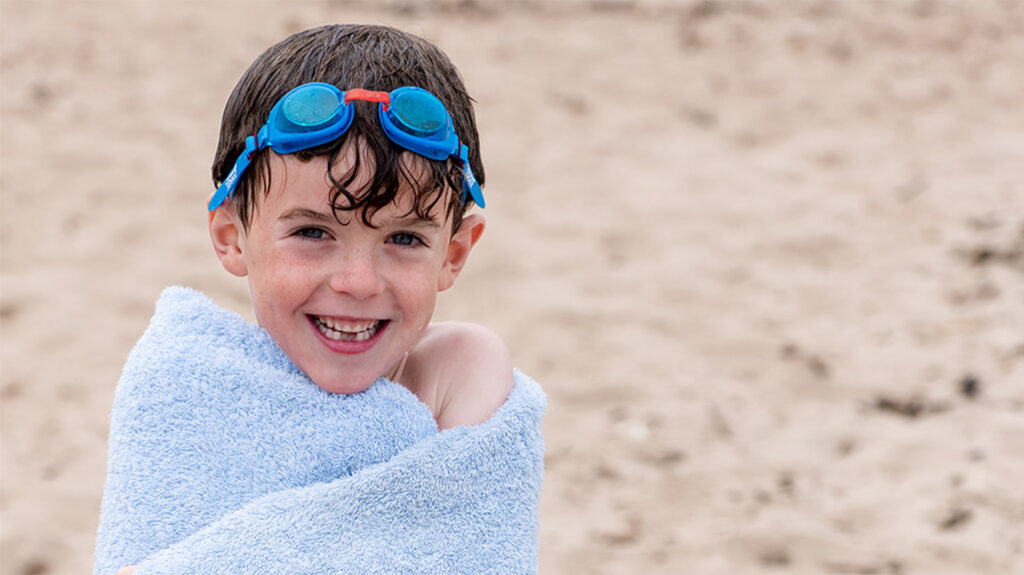 Six-year-old Bertie stands on a beach wrapped in a towel and wearing goggles on his forehead, while smiling broadly.