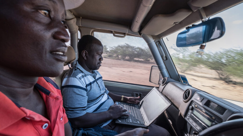 Samson sits in the passenger seat of a car using a laptop. A man sitting next to him is driving. They are travelling through a desert landscape.