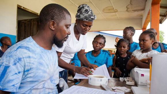 A man sits at a table with research forms in front of him, while a woman standing next to him shows him some documents. A group of primary school children wait by the table watching.