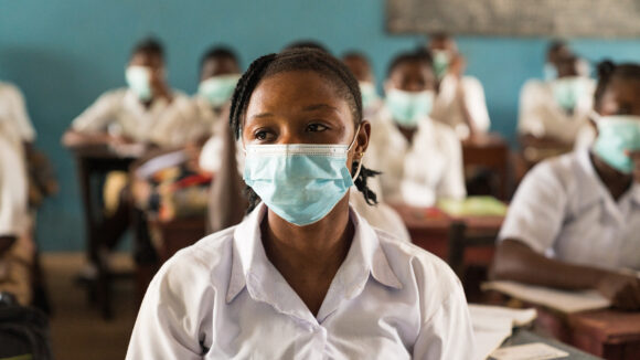 Saio sits in a classroom wearing a surgical masks. Behind her, there are rows of desks with other students.