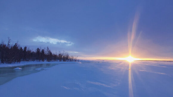 The sun rises on the horizon over a snowy expanse. On the left, there are bare trees and a river with floating ice chunks.