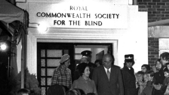 The Queen stands outside the Royal Commonwealth Society for the Blind offices while a crowd looks on.