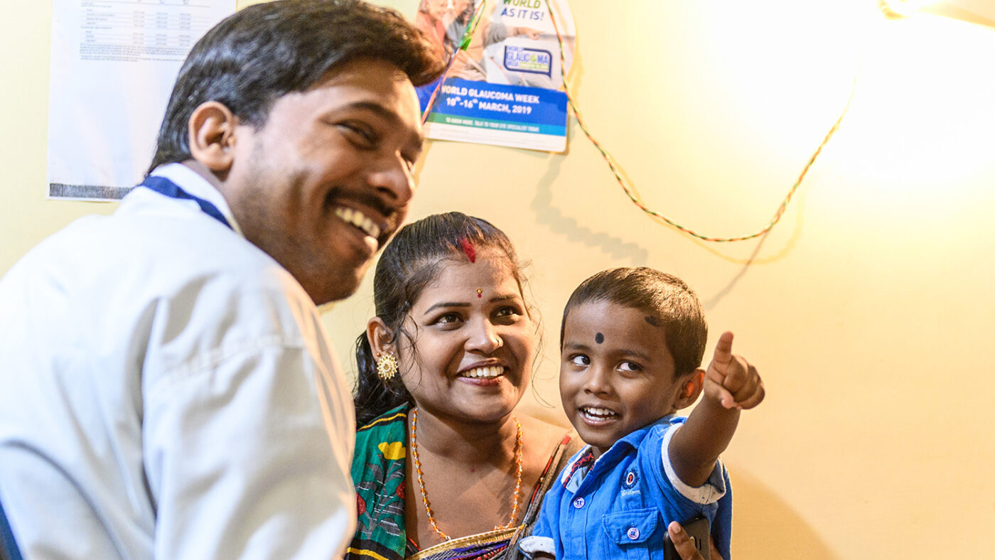 Omm smiles and points as his eyes are examined by a doctor. He's sitting on his mother's lap.