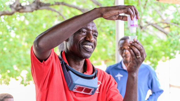 Jeremiah, an ophthalmic nurse, holds up a vial of medication. He's smiling and wearing specialist eye health equipment around his neck.