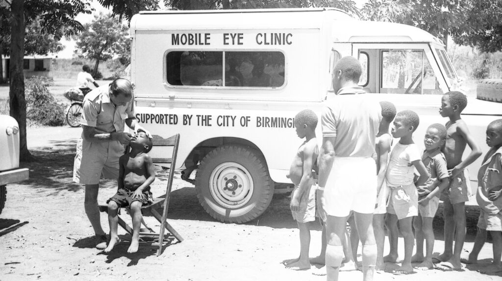 A black and white image of children having their eyes checked, in front of a van labelled Mobile Eye Unit.