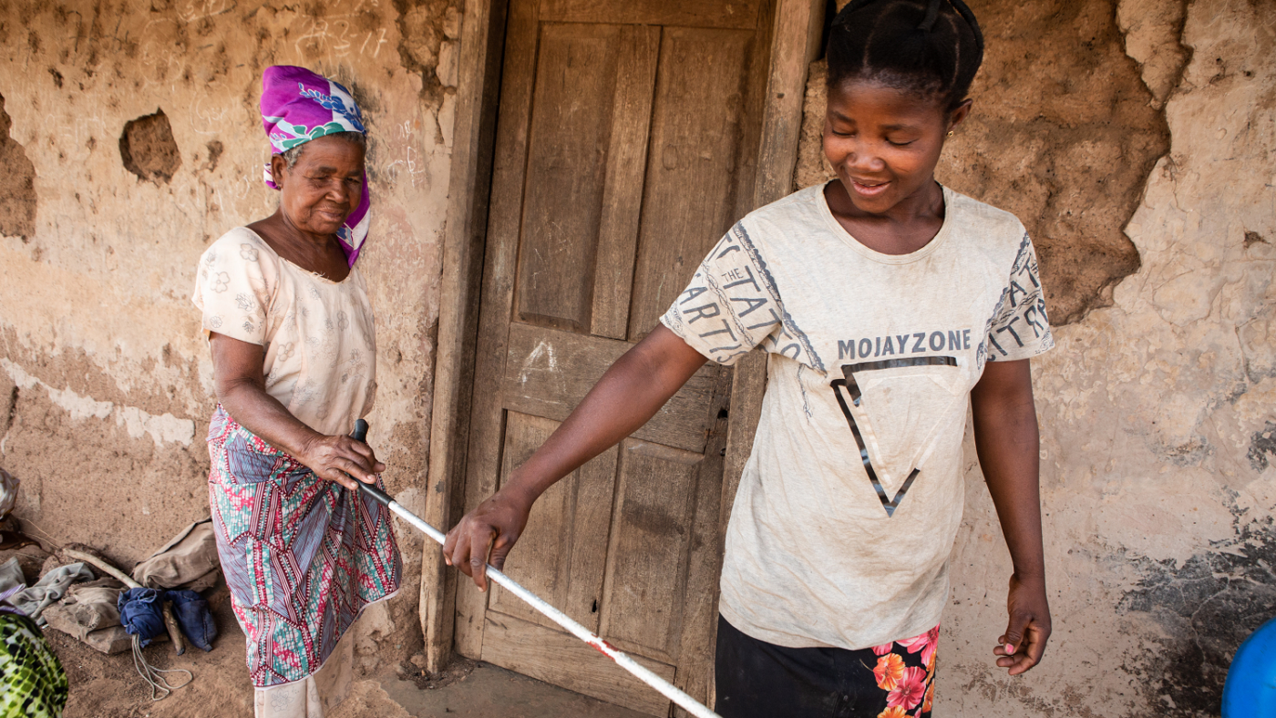 A young woman, Effia, holds a cane to lead her blind mother, Abena, outside.