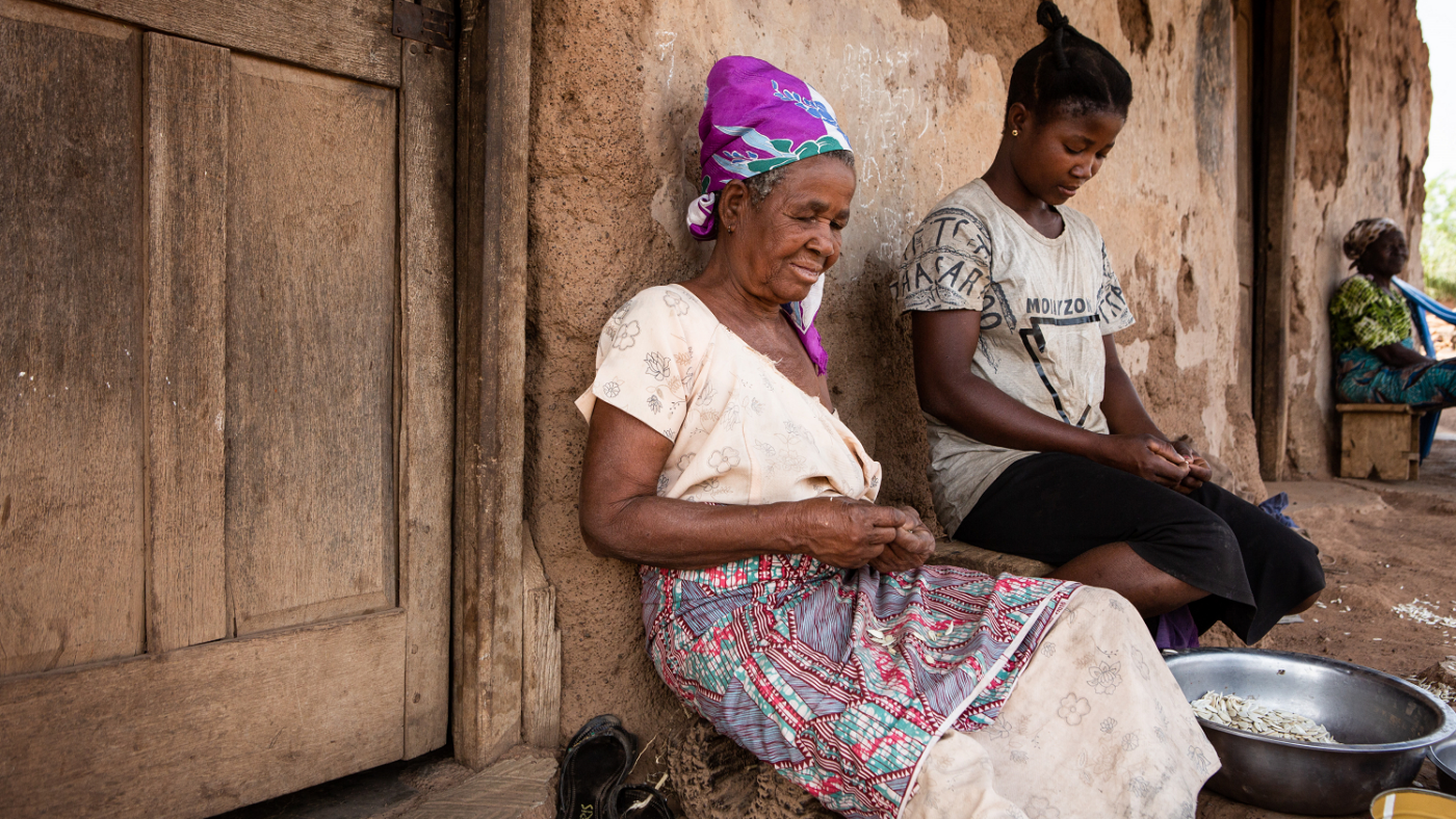 A young woman sits outside next to an older woman preparing food.