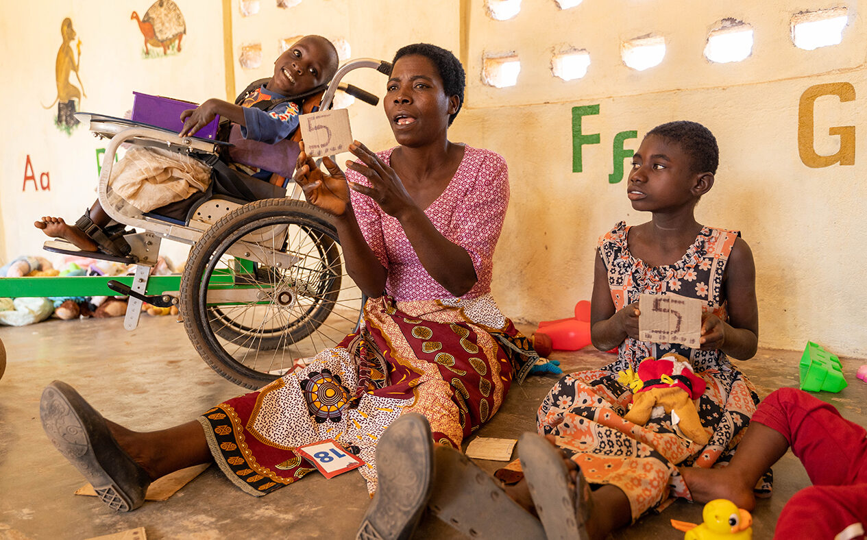 Nora teaches numbers to pre-school children. A child using a wheelchair sits on her left.