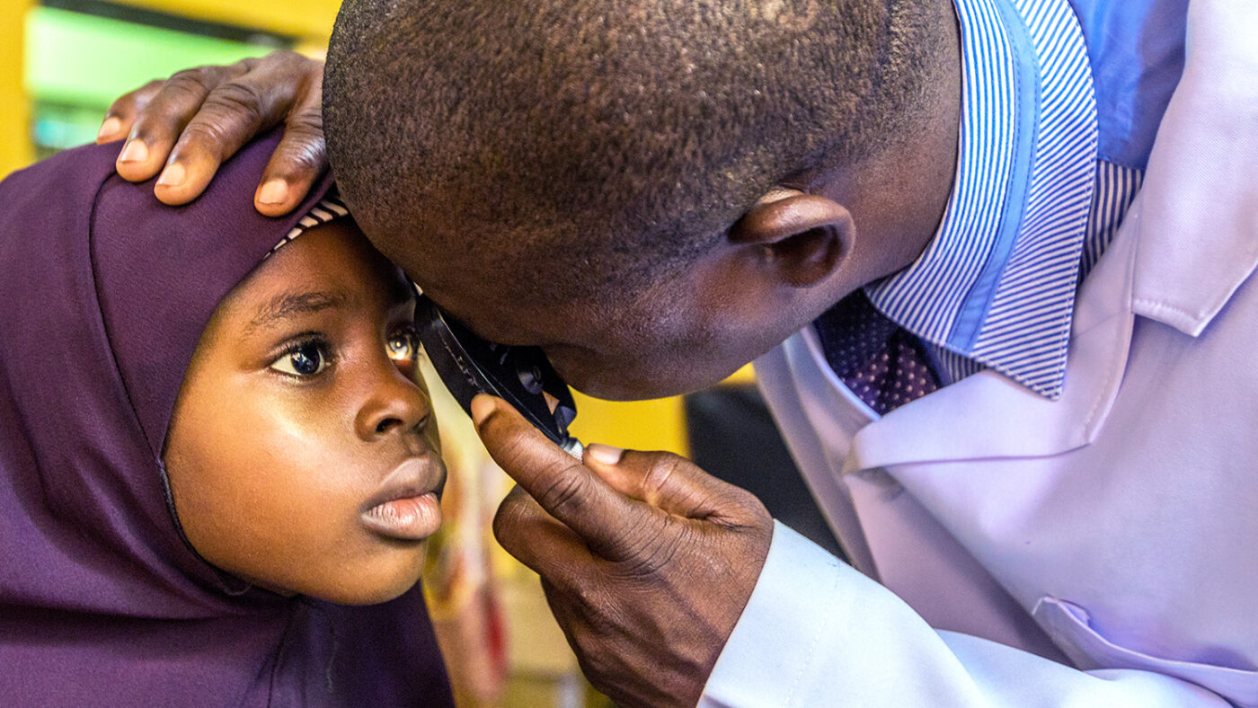 A young girl wearing a purple headscarf has her eyes examined by an ophthalmologist wearing a white coat.