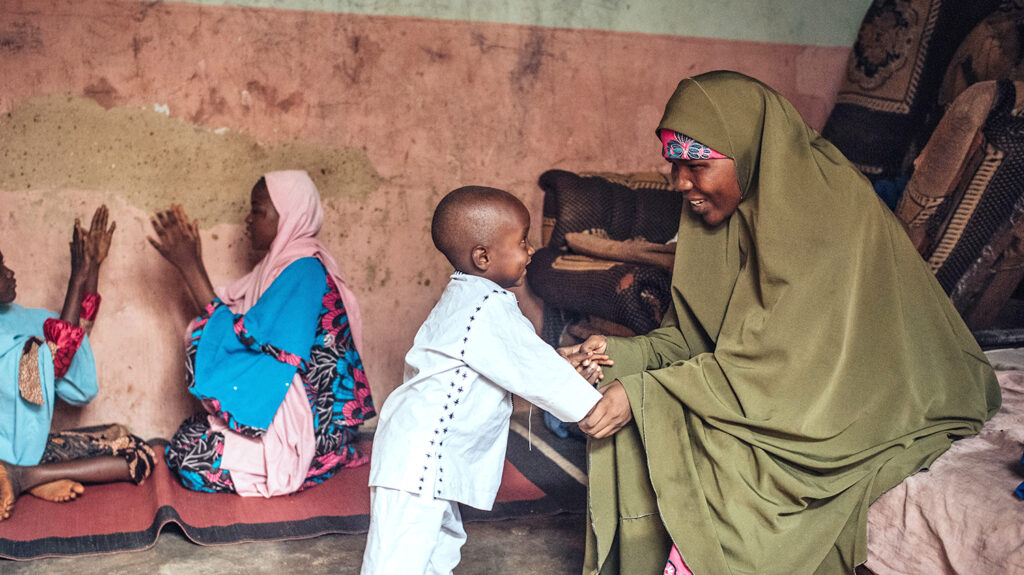 Amina, wearing a long green headscarf, smiles as she plays with her young son at their home.