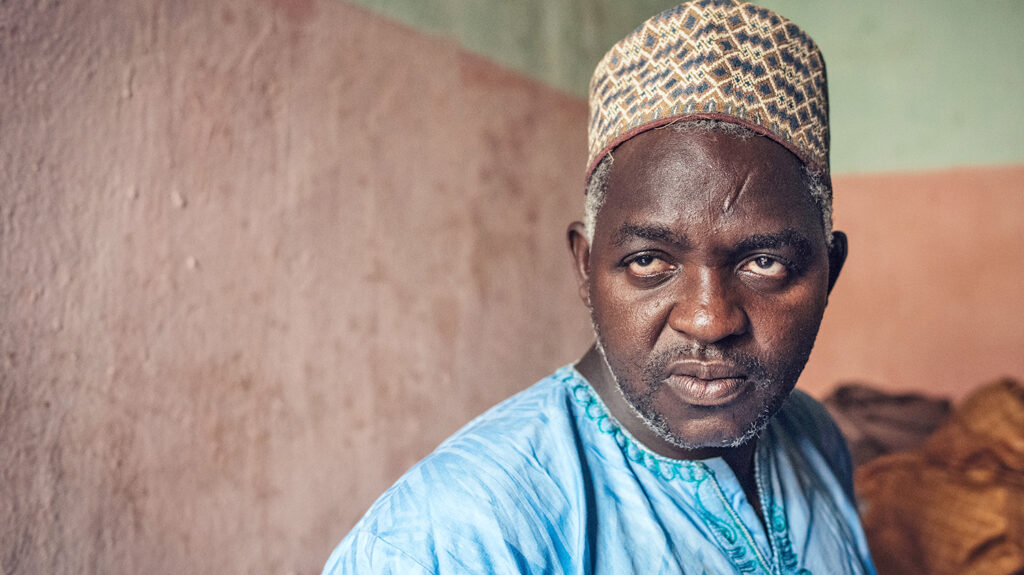 Ashafa, looking serious, wears a traditional Nigerian hat and vibrant blue shirt.