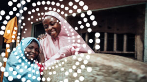 Asmau and Fatima, both wearing coloured headscarves, smile and embrace at their home in Nigeria.