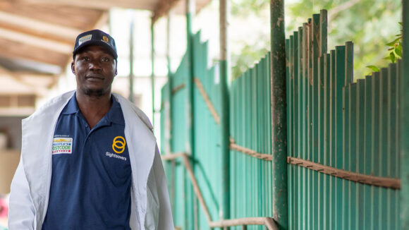 Optometrist George stands outside wearing a People's Postcode Lottery branded baseball cap and polo shirt.