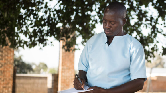 Gervazio writes a letter while sitting under a tree.