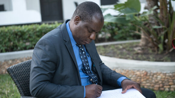 Bright sits in a chair outdoors and writes a letter. He's wearing a smart suit and tie.