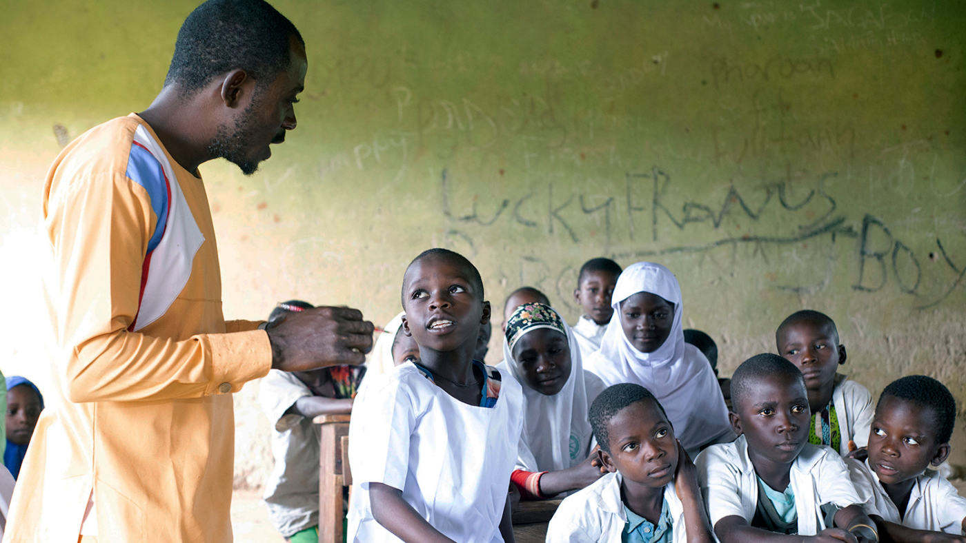 In a classroom, a teacher stands next to a student who is answering a question.