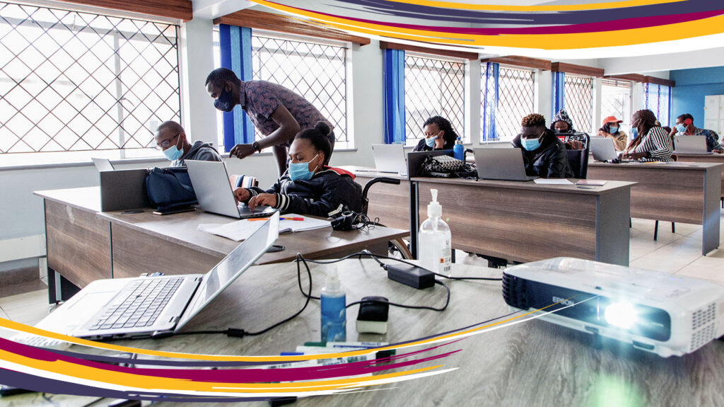 Students sit in front of laptops at desks in a classroom. In the foreground, a teacher stands next to a student, giving instruction.