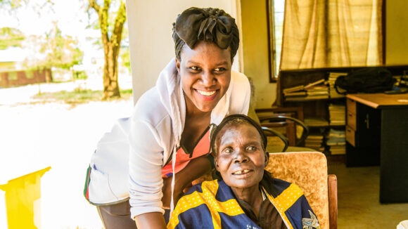 Gladys smiles while standing over an elderly woman who is sitting down.