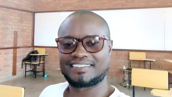 Alinafe smiles while sitting in a classroom with desks behind him.