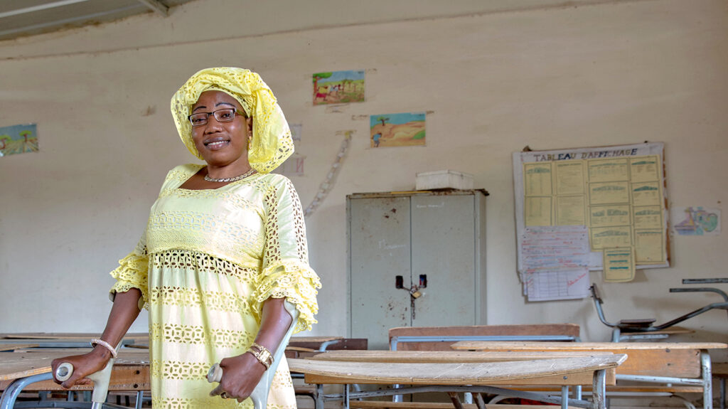 Isseu stands in a classroom holding her crutches. She's wearing a vibrant yellow dress and headscarf.
