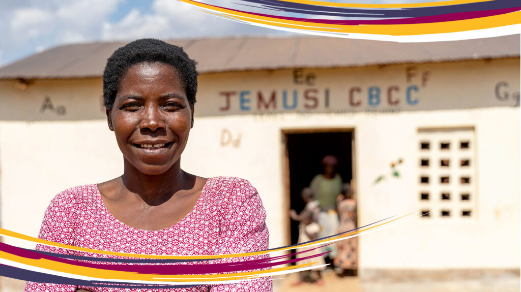 Nora stands outside the pre school where she teaches in Malawi, with her arms crossed across her chest.