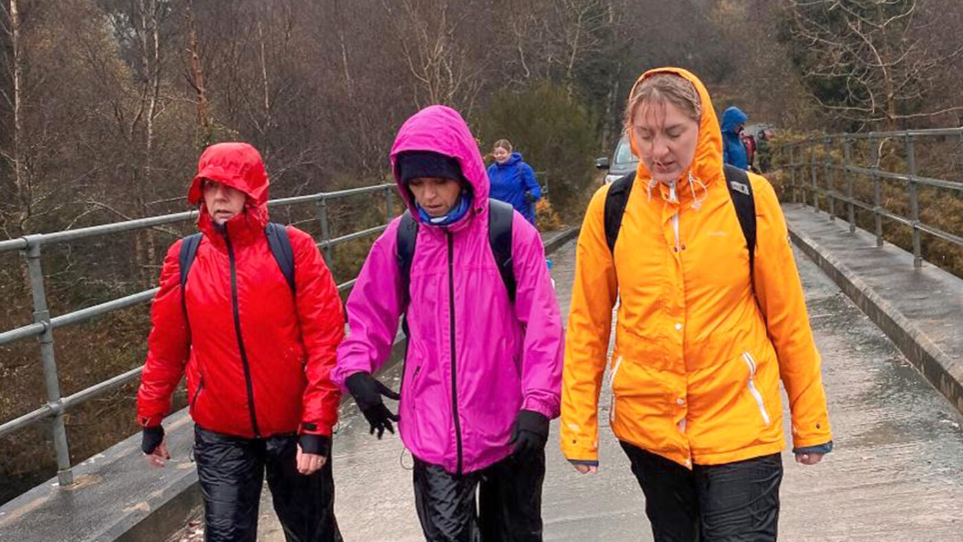 Three women wearing brightly coloured outdoor jackets hike in the wet Wicklow Mountains.