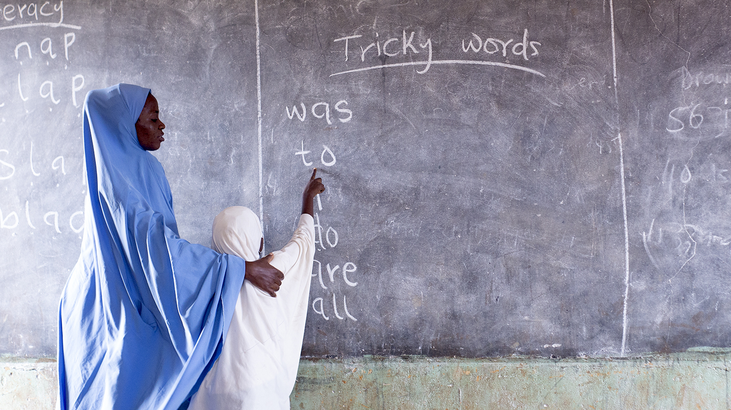 A teacher in Nigeria outs her arm on a student's shoulders as they stand at the blackboard.