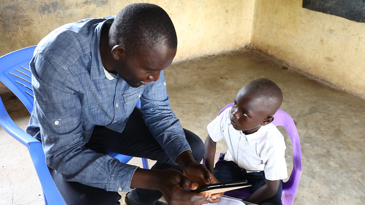 A male researcher using a tablet sits next to a young child during an early years development assessment in Kenya.