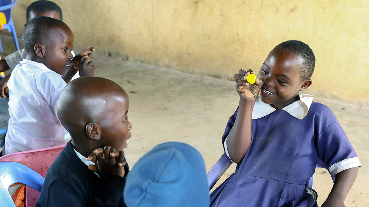 A young girl surrounded by classmates smiles while playing with a plastic toy.