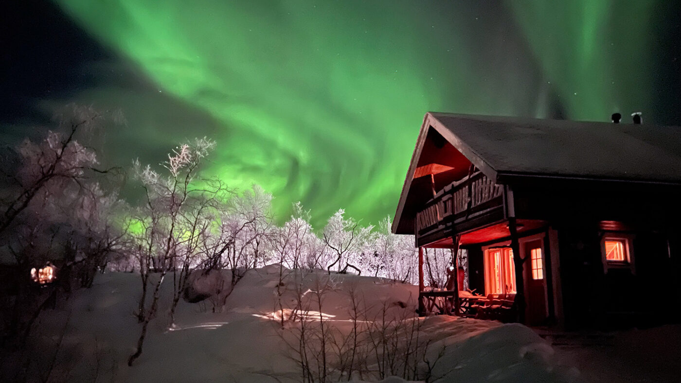 The northern lights dance behind a wooden cabin in a snowy Arctic landscape.