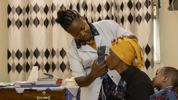 An eye doctor examines the eyes of a mother who is carrying her child on her back.