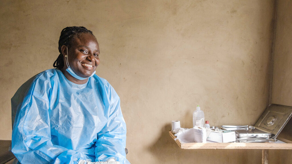 Olubunmi, a gynaecologist from Nigeria, smiles while wearing a blue plastic medical apron and mask under her chin.