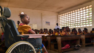8-year-old Lesline, who uses a wheelchair, sits in the corner of her classroom in Cameroon looking at the board. Her classmates sit in rows behind her.