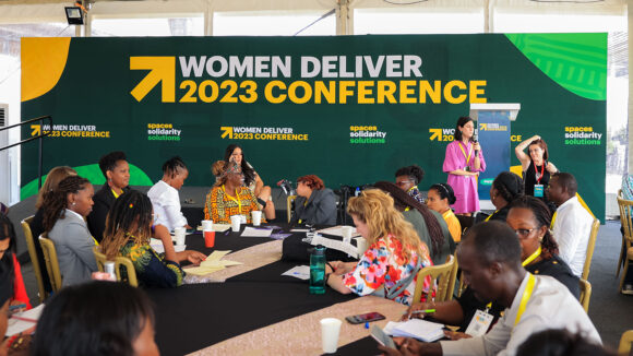 A group of people sitting at tables in front of a banner saying 'Women Deliver 2023 Conference'.