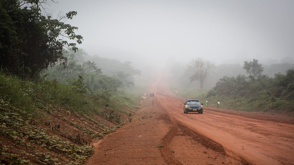 A car travels along a wide, dusty road in rural Ghana.
