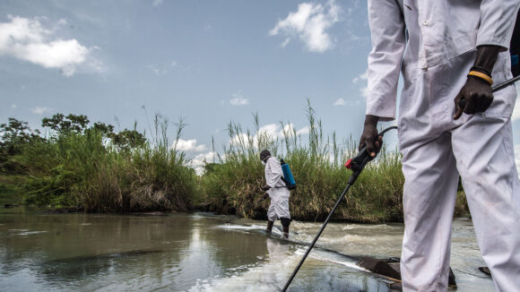 Two men wearing scrubs spray larvicide into a river in Uganda to control river blindness.