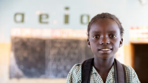 Ruth, a young student, smiles at the camera. She's wearing her green checked school shirt and carrying a backpack.
