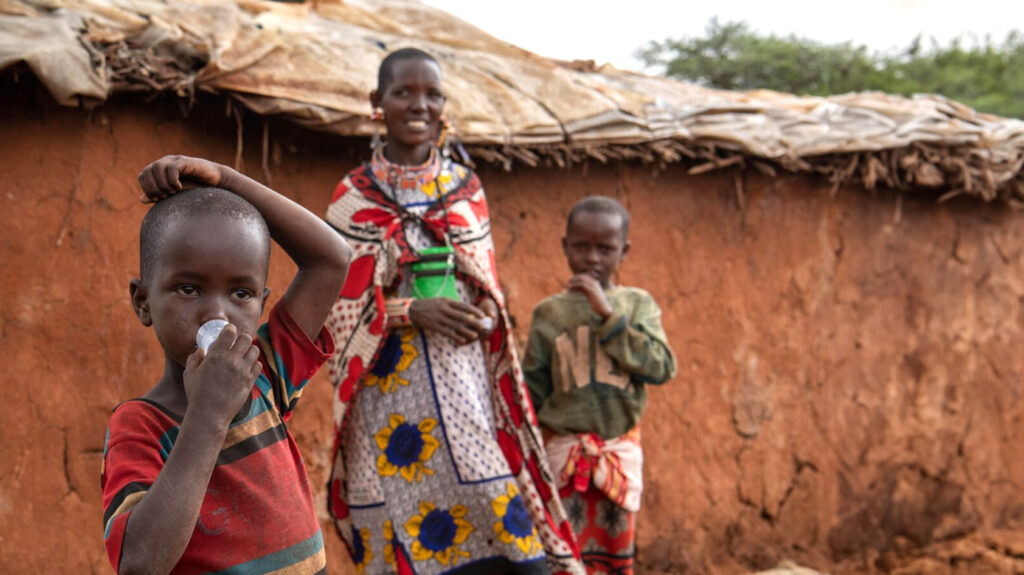 Lanoi stands outside with two people standing behind her. She takes medicine from a small cup.