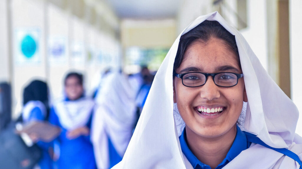 School student Zahra in the corridor at school. She's smiling and wearing a white headscarf and bright blue shirt.