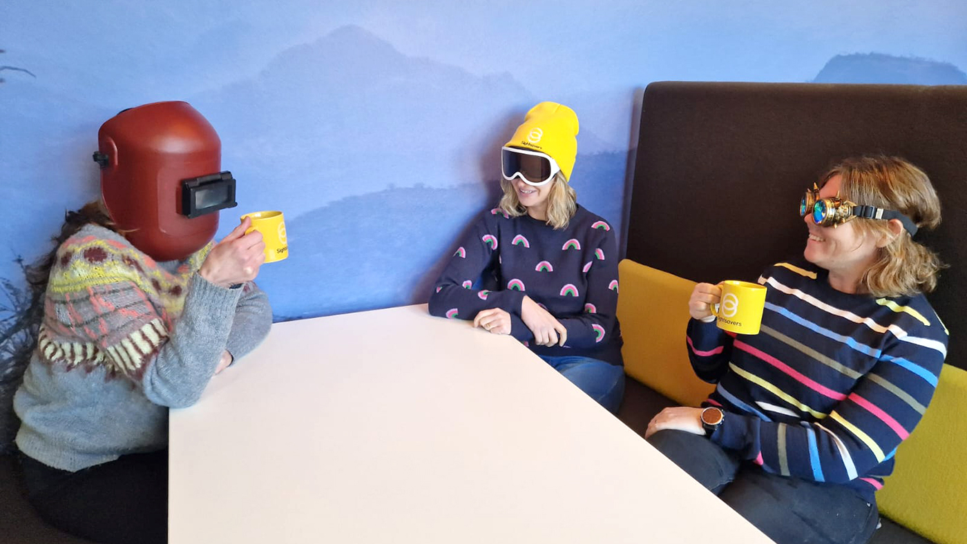 Three people sitting at a table wearing different eyewear - a welding mask, ski goggles and old-fashioned flying goggles.
