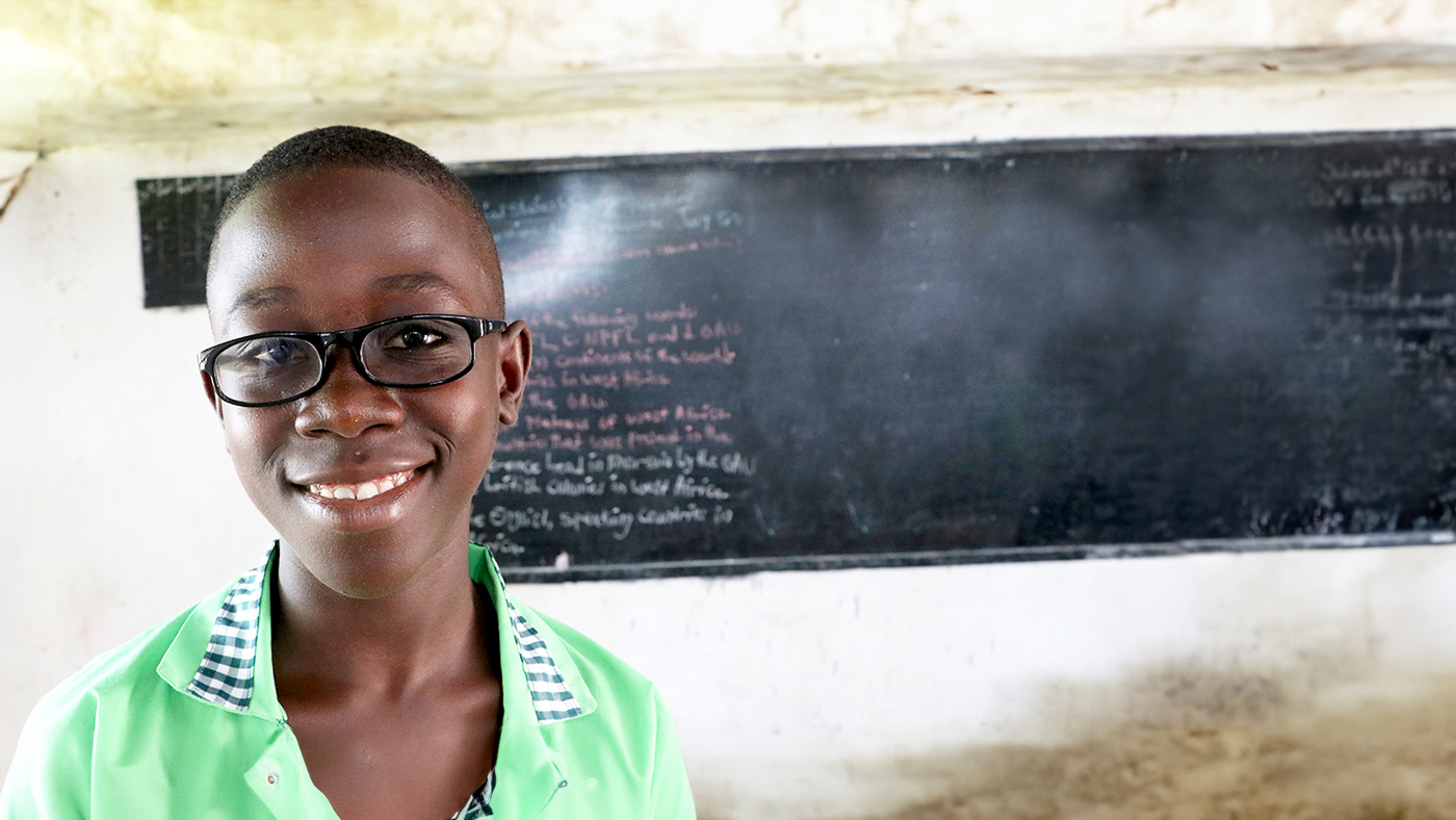 Arthur smiles int he classroom while wearing his new glasses. There's a blackboard in the background.