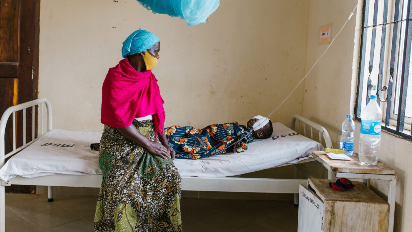 Mohamed lies on a hospital bed with bandages over his eyes. His grandmother, Sophia, sits with him.