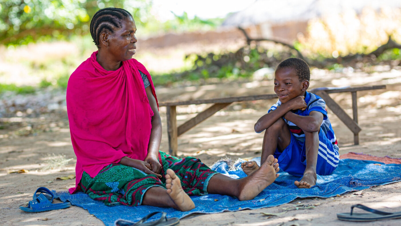 Mohamed sits outside on a blanket with his grandmother, Sophia. They are smiling.