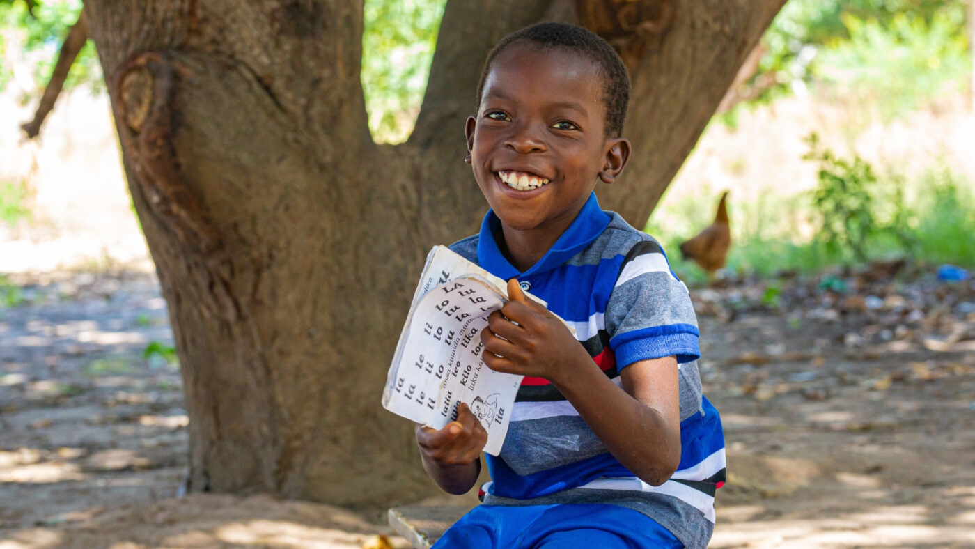 Mohamed smiles while holding a school literacy workbook. His eyes are open.