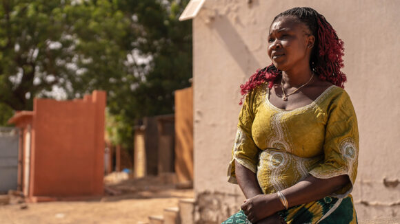 Batawouni is sitting outside a building looking into the distance. She is wearing a yellow top, gold necklace a traditional African green wraparound.