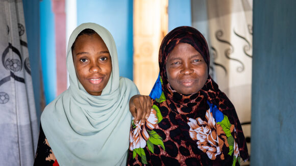 Mzowele and her daughter sit together at home in their village. They are both wearing hijabs.
